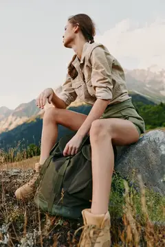 Молодая женщина в рубашке цвета хаки и шортах сидит на камне с рюкзаком в горной природе