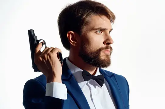 Бородатый мужчина в смокинге держит пистолет у плеча на белом фоне