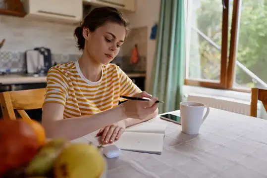 Женщина за кухонным столом дома, работает с планшетом и блокнотом, мягкий солнечный свет, непринуждённая поза, организованное пространство, продуктивное настроение, творческая сосредоточенность…
