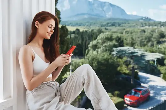 Рыжеволосая девушка с телефоном у окна на фоне гор