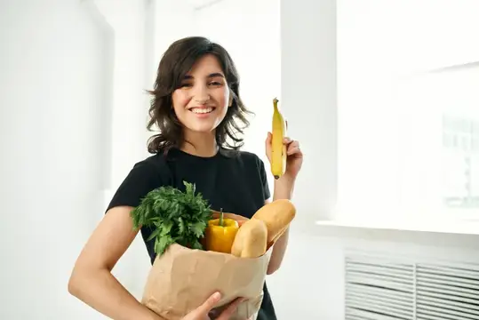 Улыбающаяся брюнетка с пакетом продуктов и бананом в руке