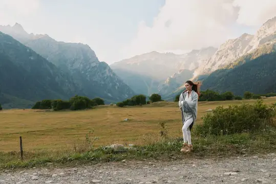 Ее развевающиеся на ветру волосы и повседневная куртка ловят дуновение ветра, когда она смотрит в сторону гор, создавая тихий момент на открытом воздухе с естественным светом и умиротворенным…