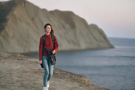 Улыбающаяся девушка с рюкзаком гуляет по берегу у скалы
