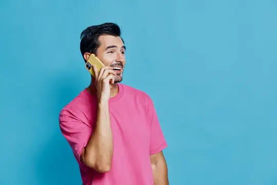 Улыбающийся темноволосый мужчина в розовой футболке говорит по телефону на синем фоне