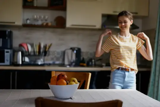 Улыбающаяся девушка танцует на кухне рядом с вазой фруктов