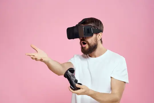 Бородатый мужчина в VR-очках с геймпадом удивлённо жестикулирует на розовом фоне