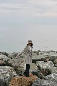 Женщина в плаще и кепке стоит на камнях у моря в пасмурный день