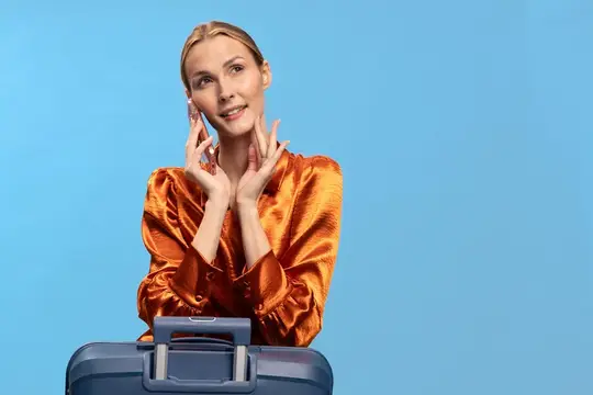 Улыбающаяся блондинка с чемоданом говорит по телефону на голубом фоне