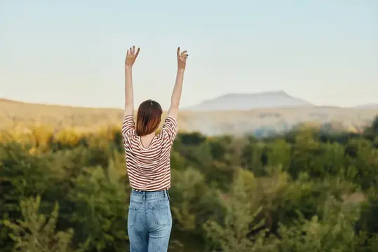 Девушка с поднятыми руками стоит спиной на фоне гор и леса