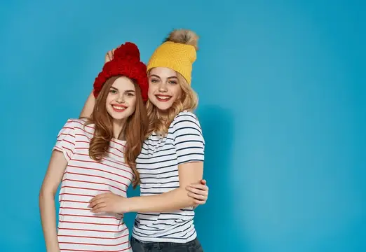 Две улыбающиеся девушки в вязаных шапках обнимаются на синем фоне