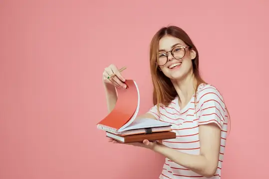 Молодая студентка с книгами на розовом фоне, в очках, крупный план лица