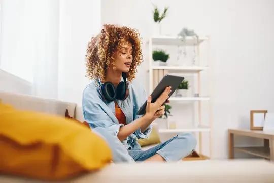 Счастливая женщина наслаждается онлайн-игрой на диване в уютной гостиной с планшетом и наушниками