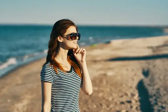 Женщина в футболке и солнцезащитных очках стоит на песчаном берегу пляжа у моря
