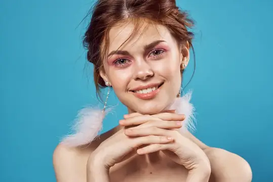 Женщина с обнажёнными плечами, пушистыми серьгами и ярким макияжем на синем фоне