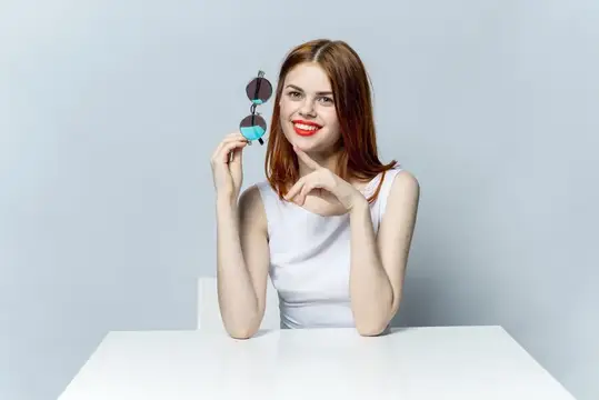 На высококачественной фотографии изображена симпатичная женщина, сидящая за белым столом с бокалами в руках и улыбающаяся