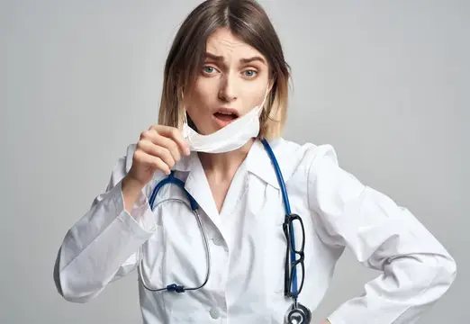 Удивлённая женщина-врач приспускает медицинскую маску на сером фоне
