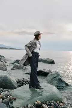 Женщина в плаще и кепке стоит на камнях у моря