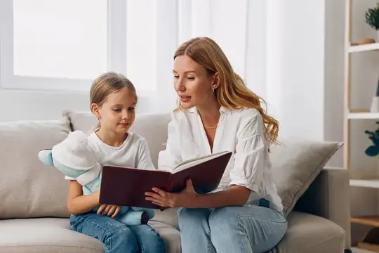 Светловолосая мама читает книгу дочке на диване