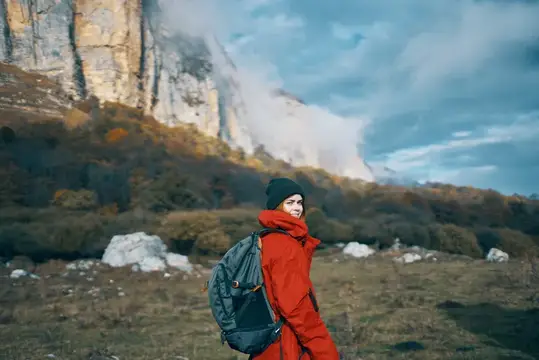 Женщина с рюкзаком в красной куртке путешествует на фоне голубого неба, высоких гор и скалистого ландшафта