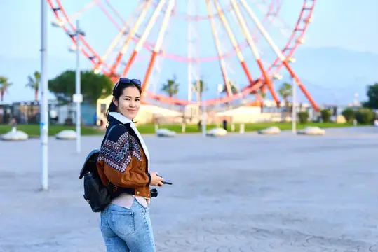 Уличная сцена: женщина в повседневной куртке и с рюкзаком держит фотоаппарат рядом с колесом обозрения