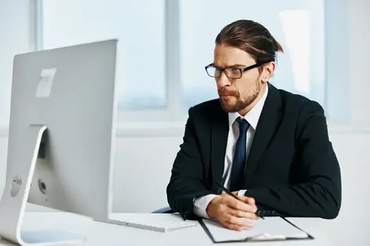 Бородатый мужчина в очках и костюме работает за компьютером в офисе
