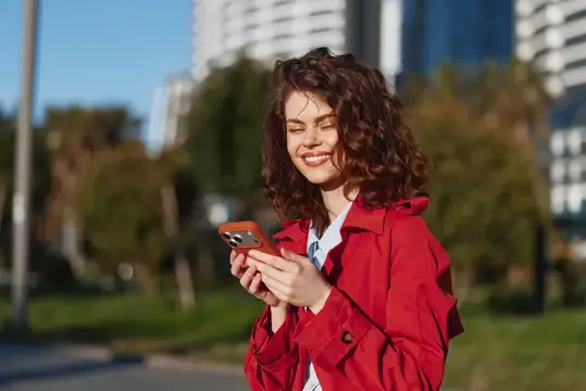 Улыбающаяся кудрявая девушка в красном плаще смотрит в телефон на улице