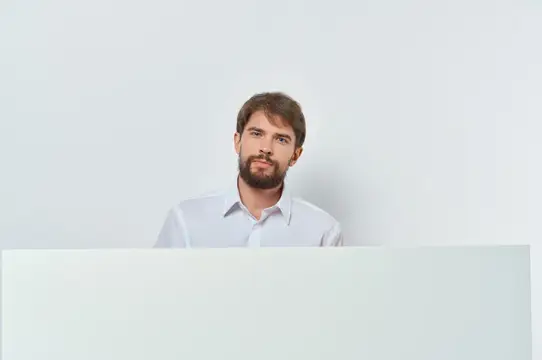 Молодой бородатый мужчина в белой рубашке выглядывает из-за белой панели