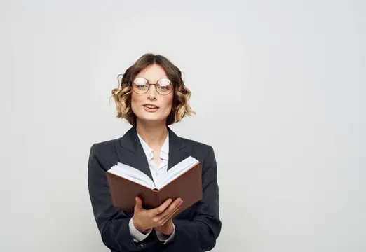 Молодая женщина в очках и деловом костюме читает книгу на светлом фоне