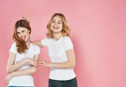 Две веселые девушки в белых футболках дурачатся на розовом фоне