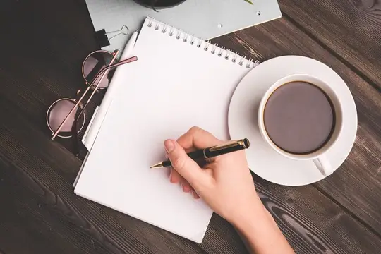 Рука с ручкой над блокнотом рядом с чашкой кофе и очками