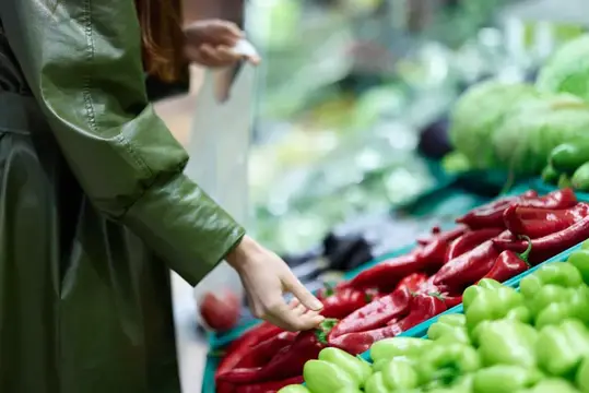 Женщина в зелёном плаще выбирает красный перец на рынке