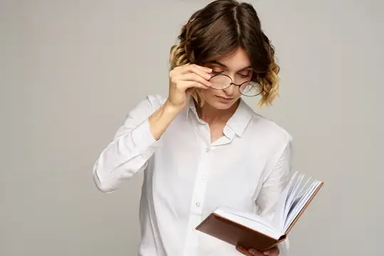 Уставшая женщина в белой рубашке поправляет очки и читает книгу