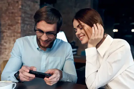 Мужчина и женщина смеются, глядя в смартфон в кафе