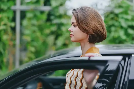 Молодая женщина с каре смотрит вдаль, стоя у чёрной машины