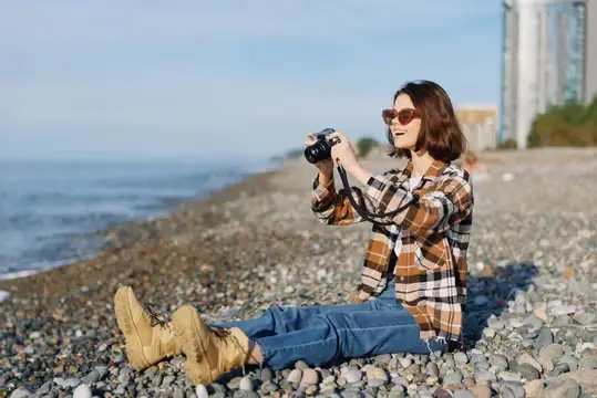 Девушка в клетчатой рубашке фотографирует на каменистом берегу