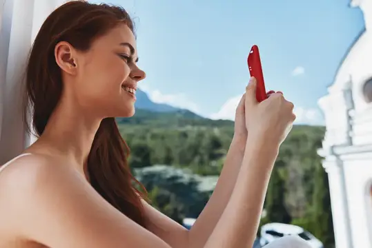 Улыбающаяся шатенка фотографирует пейзаж на красный смартфон у окна