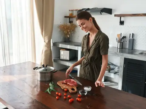 Улыбающаяся девушка режет мясо на деревянной доске на кухне