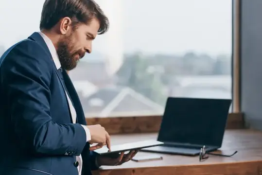 Бородатый мужчина в костюме работает с планшетом у окна