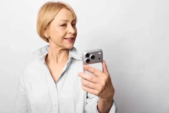 Улыбающаяся блондинка в белой рубашке смотрит в смартфон на светлом фоне