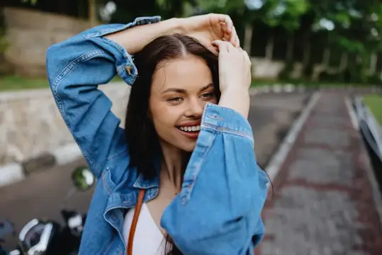 Улыбающаяся темноволосая девушка в джинсовой куртке поправляет волосы на улице
