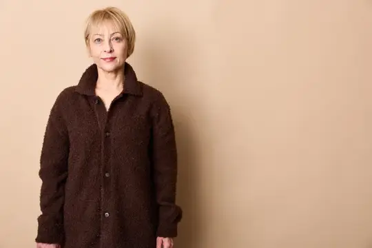 Портрет пожилой женщины, стоящей в студийном свете, бежевый фон, тёмная застёгнутая куртка, задумчивое спокойное лицо, нейтральный взгляд, удобная поза