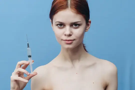 Рыжеволосая девушка с обнажёнными плечами держит шприц на голубом фоне
