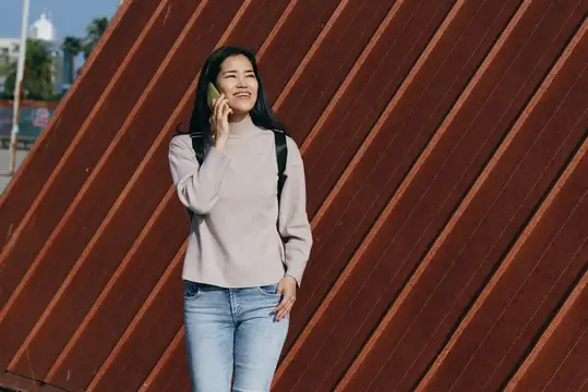 Улыбающаяся азиатская девушка говорит по телефону на фоне коричневой стены
