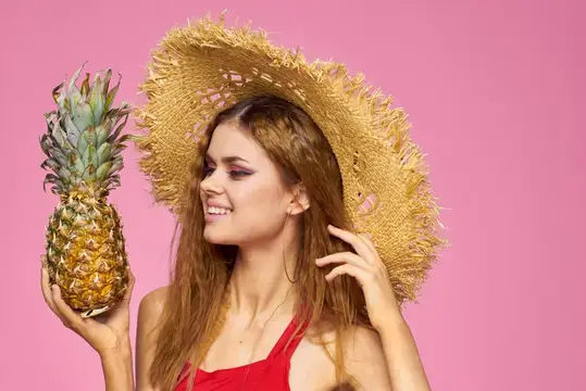 Улыбающаяся девушка в соломенной шляпе держит ананас на розовом фоне