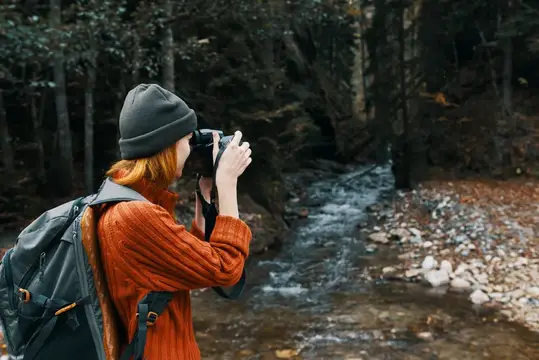 Рыжеволосая девушка с рюкзаком фотографирует лесной ручей