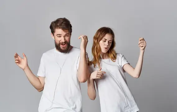 Весёлые мужчина и женщина в наушниках слушают музыку и танцуют на сером фоне