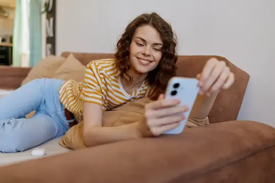 Улыбающаяся девушка лежит на диване и смотрит в смартфон