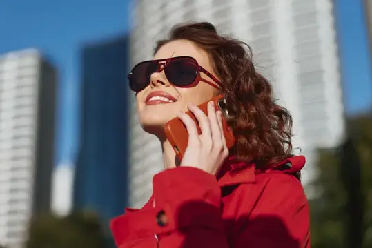 Улыбающаяся женщина в красном плаще и солнцезащитных очках говорит по телефону на фоне небоскрёбов