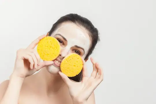Молодая женщина с глиняной маской держит жёлтые спонжи у лица