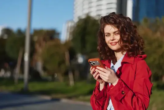 Кудрявая девушка в красном плаще смотрит на смартфон на улице